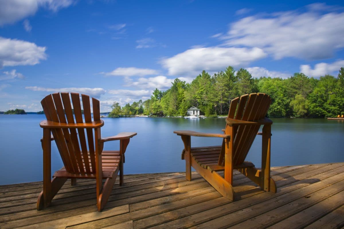 Two wooden Adirondack chairs on a dock facing a serene lake with lush green trees.